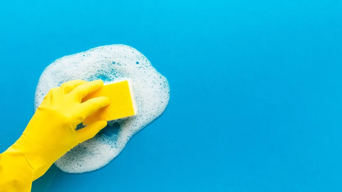 Mão com luva amarela limpando com espuma uma superfície azul (foto de banco de imagens do acervo da Uniclean)